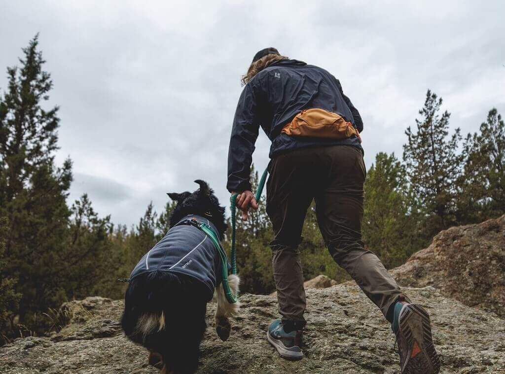 A man wearing the Ruffwear Home Trail Hip Pack and hiking with his dog on a rocky trail with trees in the background