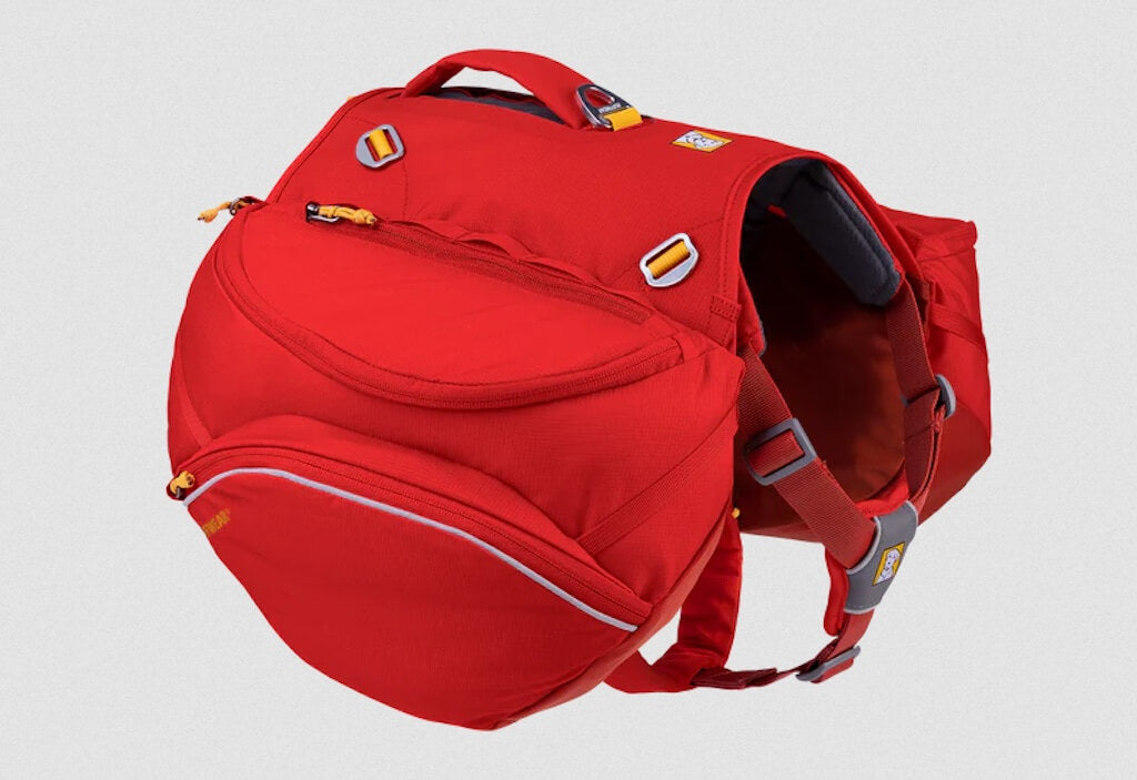 A red dog backpack called a Palisades Pack by Ruffwear with saddlebags and a padded back handle, designed for multi-day adventures.