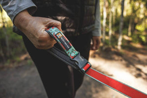 A close-up of a person's hand holding the padded handle of the Crag Leash in Tropical Bloom pattern, set against a blurred forest background.